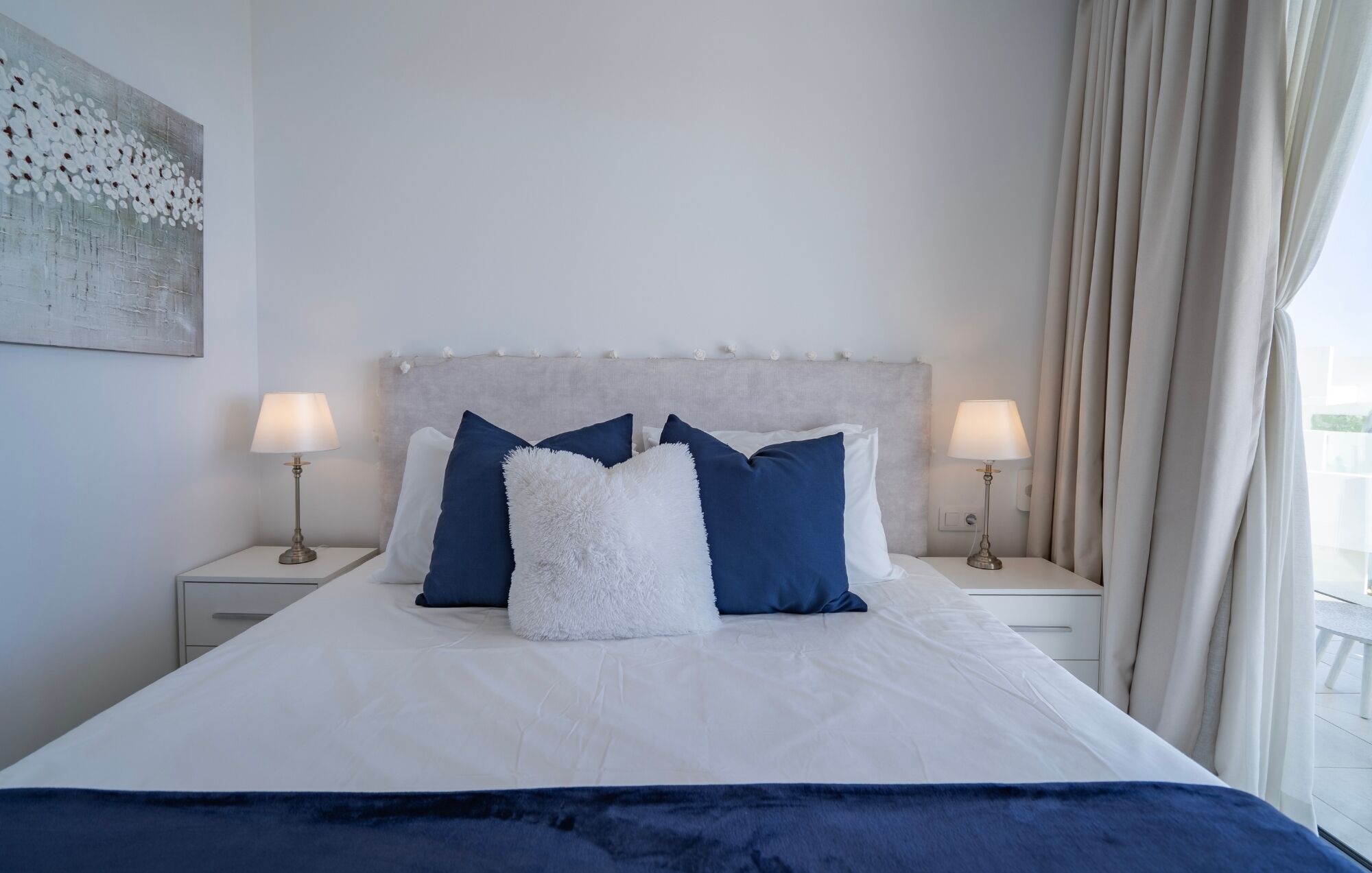 A neatly made bed with a white and blue color scheme, two bedside tables with lamps, and a large window with curtains.