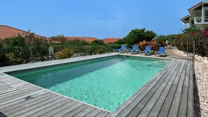 A rectangular swimming pool with a wooden deck, surrounded by lounge chairs and lush greenery. - Luxurious Apartment in Jan Thiel with Pool (Jan Thiel)