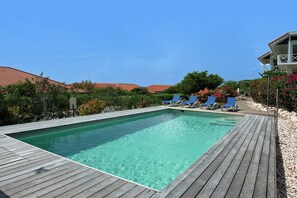 A rectangular swimming pool with a wooden deck, surrounded by lounge chairs and lush greenery. - Luxurious Apartment in Jan Thiel with Pool (Jan Thiel)