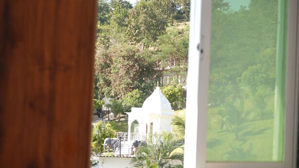 A room with a wooden headboard and a window overlooking a white building with a dome, surrounded by greenery. - Venkat's The MOUNTAIN by Redwings (Khalapur)