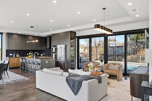 A modern kitchen with dark cabinetry, a central island, and a dining area with bar stools. - Private Pool + Firepit | Hill Views | Mins to Lake Travis | Hunters Estate (Austin)