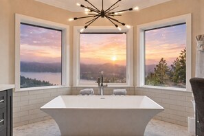 Un baño moderno con una bañera independiente, grandes ventanas que ofrecen vistas panorámicas de un atardecer sobre un lago, y un moderno accesorio de iluminación.