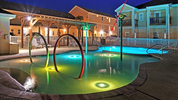 Ein Schwimmbecken mit bunten Lichtern, ein überdachter Bereich mit Stühlen und ein Gebäude im Hintergrund.