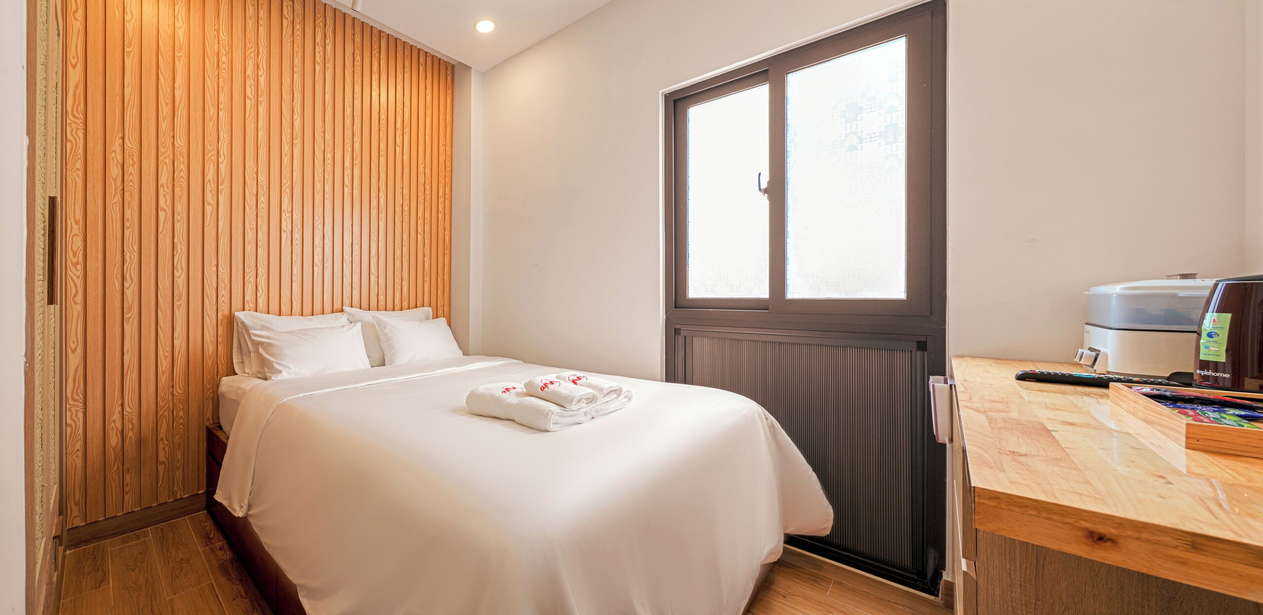 A hotel room with a wooden panel wall, a bed with white linens, a window with blinds, a wooden desk with a remote control, and a minibar.