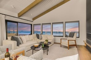A modern living room with a large sofa, a coffee table, and a TV. There are large windows with a view of the ocean and a sunset. - Oceanfront Cayucos Home + On the Sand + Firepit + Views + Steps to Cayucos Pier (Cayucos)