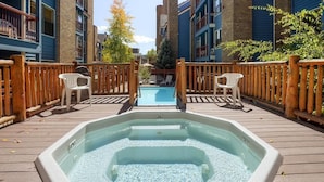 Un bain à remous sur une terrasse en bois, avec une piscine en arrière-plan, le tout entouré d’immeubles.