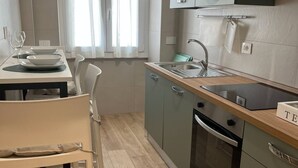 Cocina funcional con lavabo, estufa y armarios. Se ve una mesa de comedor con sillas y una ventana con cortinas.