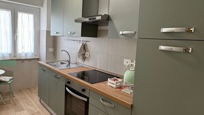 Una cocina moderna con horno empotrado, un lavamanos y una estufa. Se aprecia una ventana con cortinas, una silla verde y una mesa.