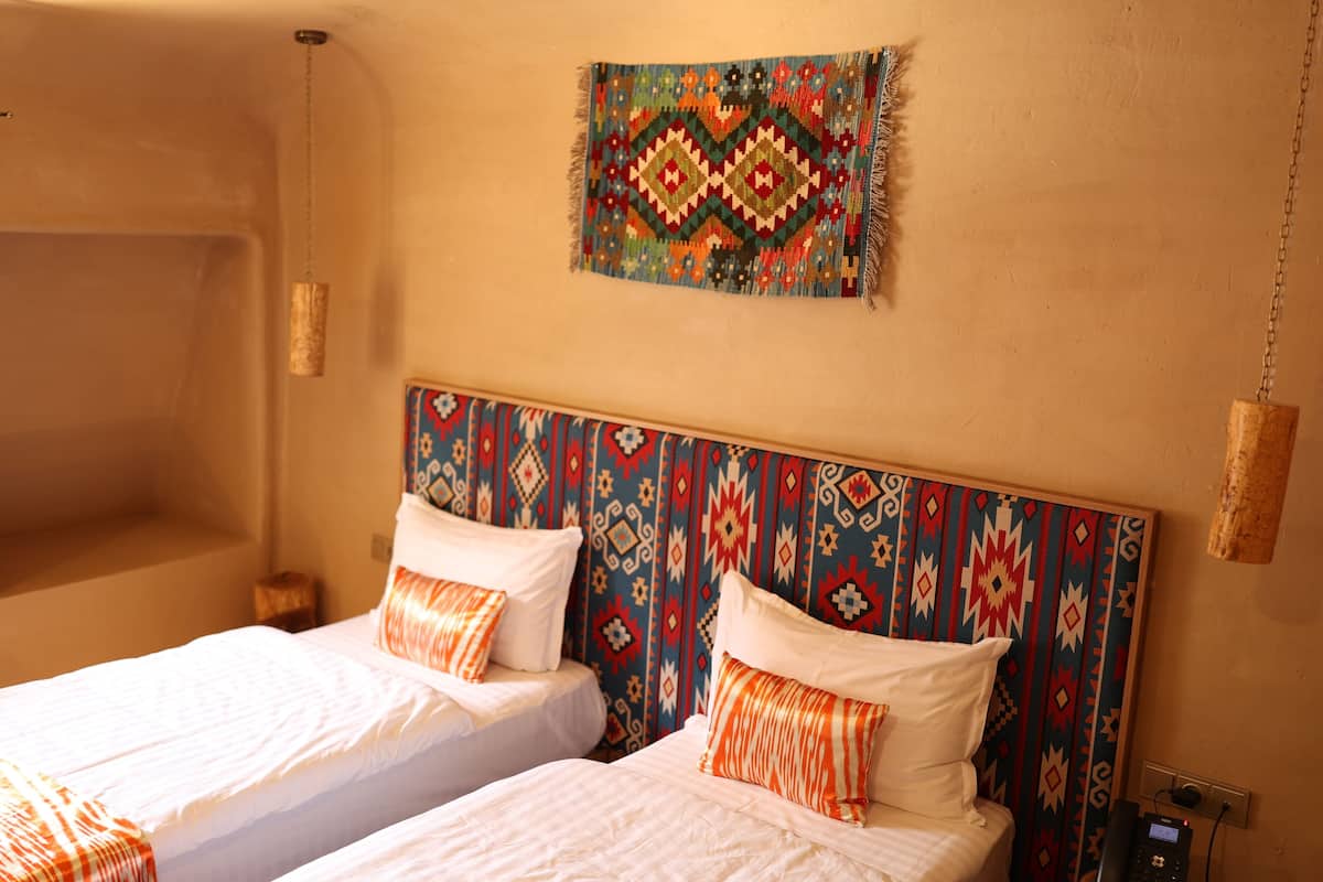 Une chambre d’hôtel avec deux lits, une tête de lit à motifs, une tapisserie murale et une table de chevet avec une lampe.
