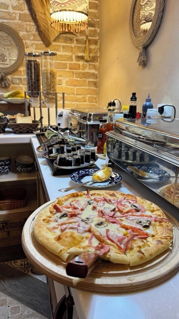 A pizza with various toppings on a wooden board, a buffet table with assorted foods, and a brick wall with a hanging light fixture. - Old Tashkent Heritage   (Tashkent)