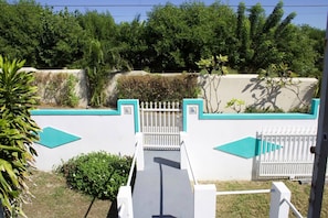A gated entrance with a white fence and turquoise accents, surrounded by lush greenery and a concrete wall.