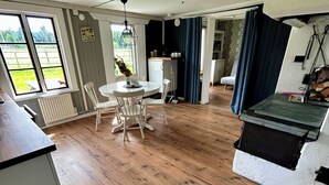 A spacious room with a wooden floor, a dining table with chairs, a small kitchen area, and a view through large windows. - Valstabacken -charming countryside cottage in peaceful Swedish nature (Möklinta)