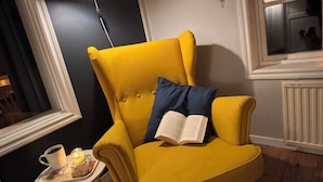 A yellow armchair with a blue pillow, a small table with a cup and a candle, and a lamp on the wall. - Valstabacken -charming countryside cottage in peaceful Swedish nature (Möklinta)