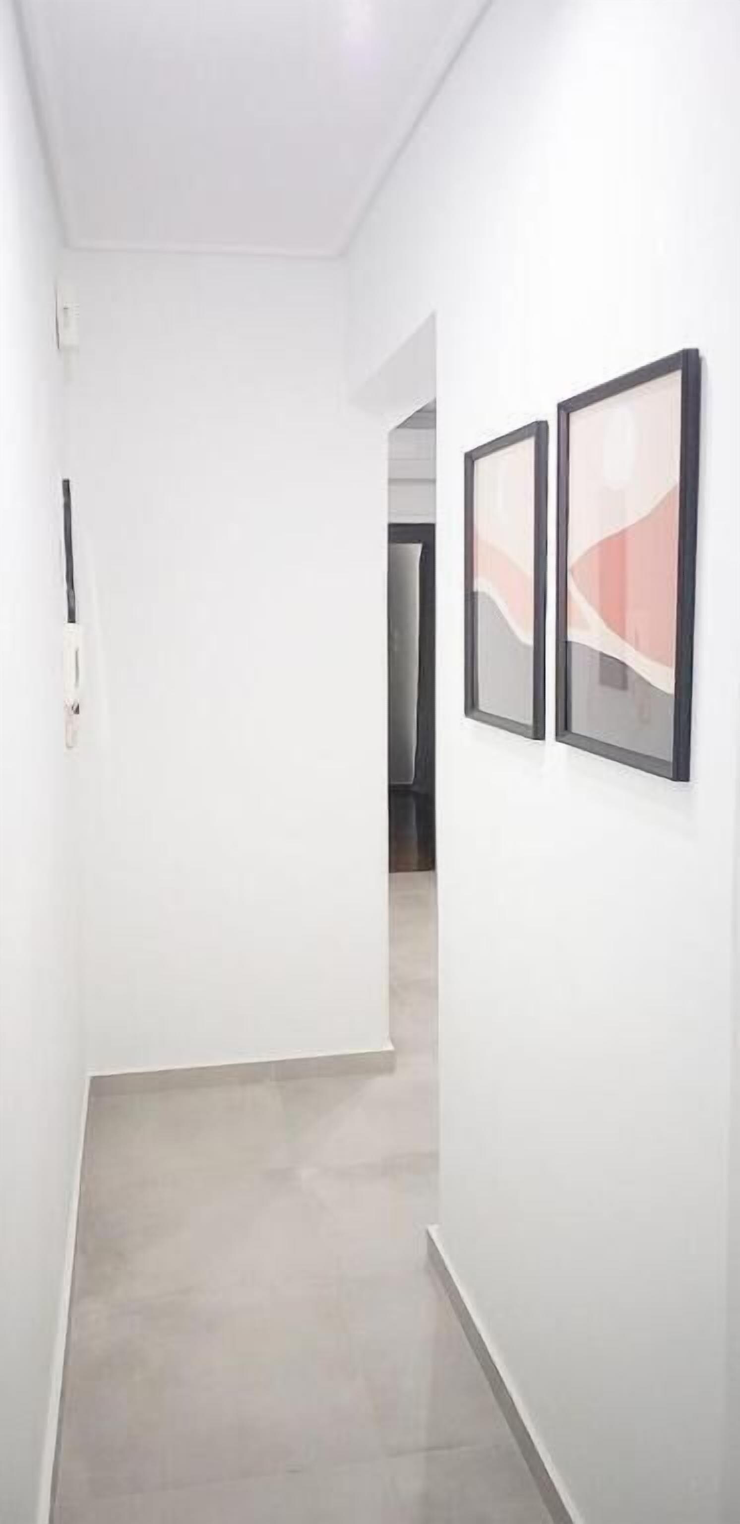 A narrow hallway with white walls and a tiled floor. There are two framed artworks on the right wall.