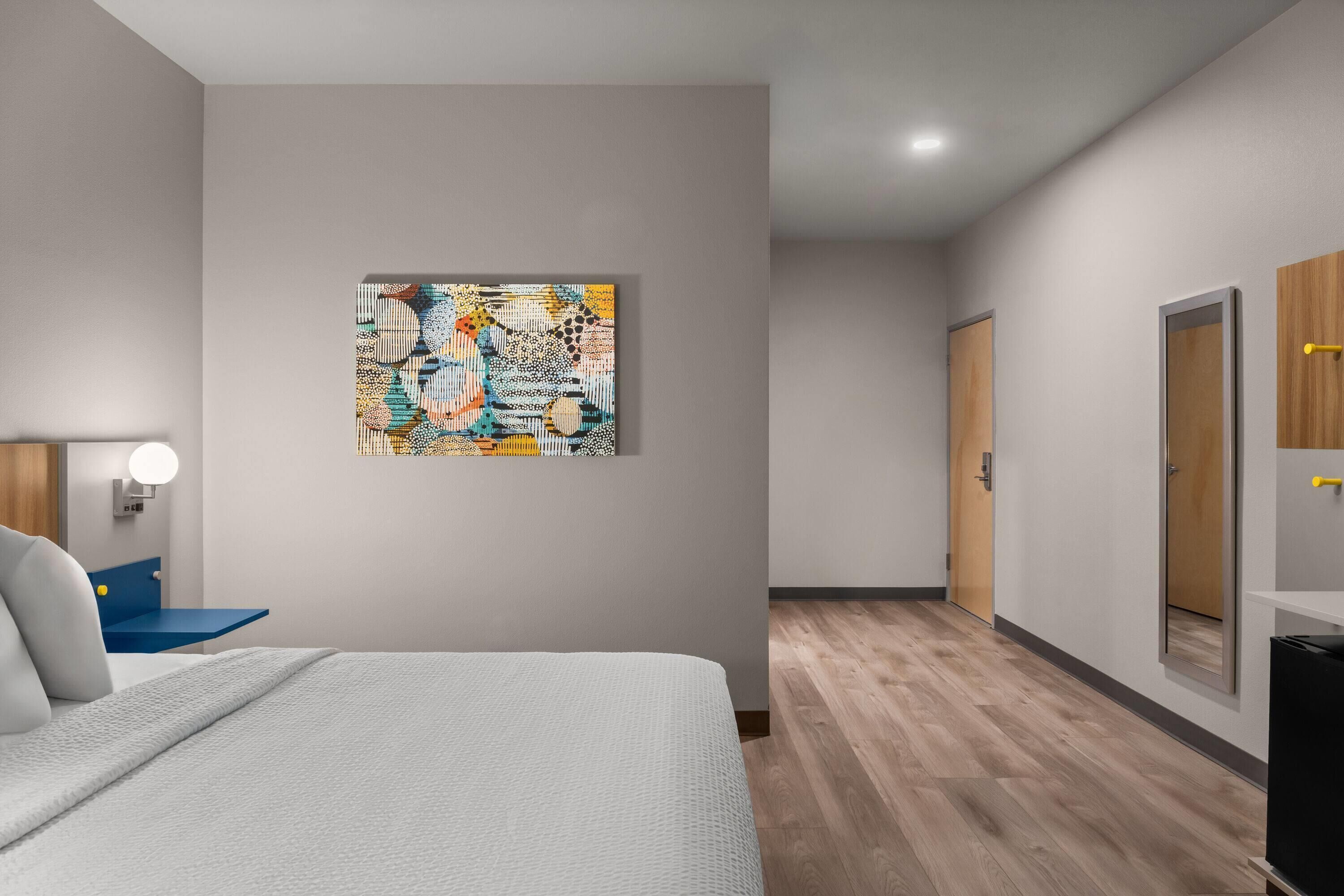 Ein modernes Hotelzimmer mit einem Bett, einem Wandbild, einem Nachttisch mit Lampe und Holzboden.