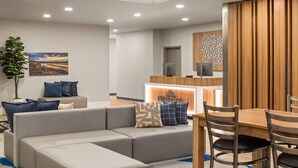 Habitación de hotel moderna con sofá modular, mesa de comedor con sillas, pequeña cocina y un ventanal grande.
