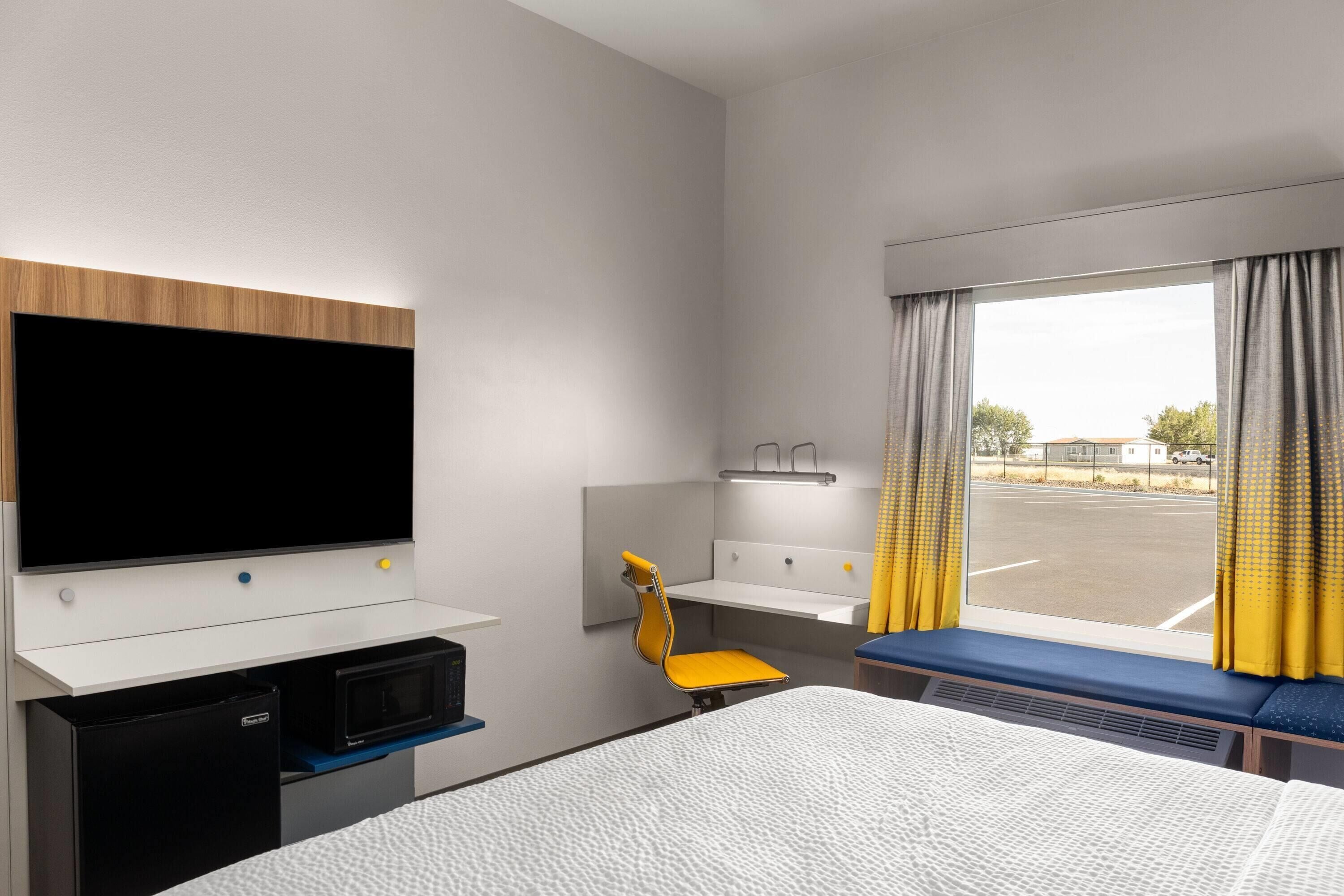A hotel room with a bed, a desk with a flat-screen TV, a microwave, a small chair, a window with curtains, and a view of the outside.