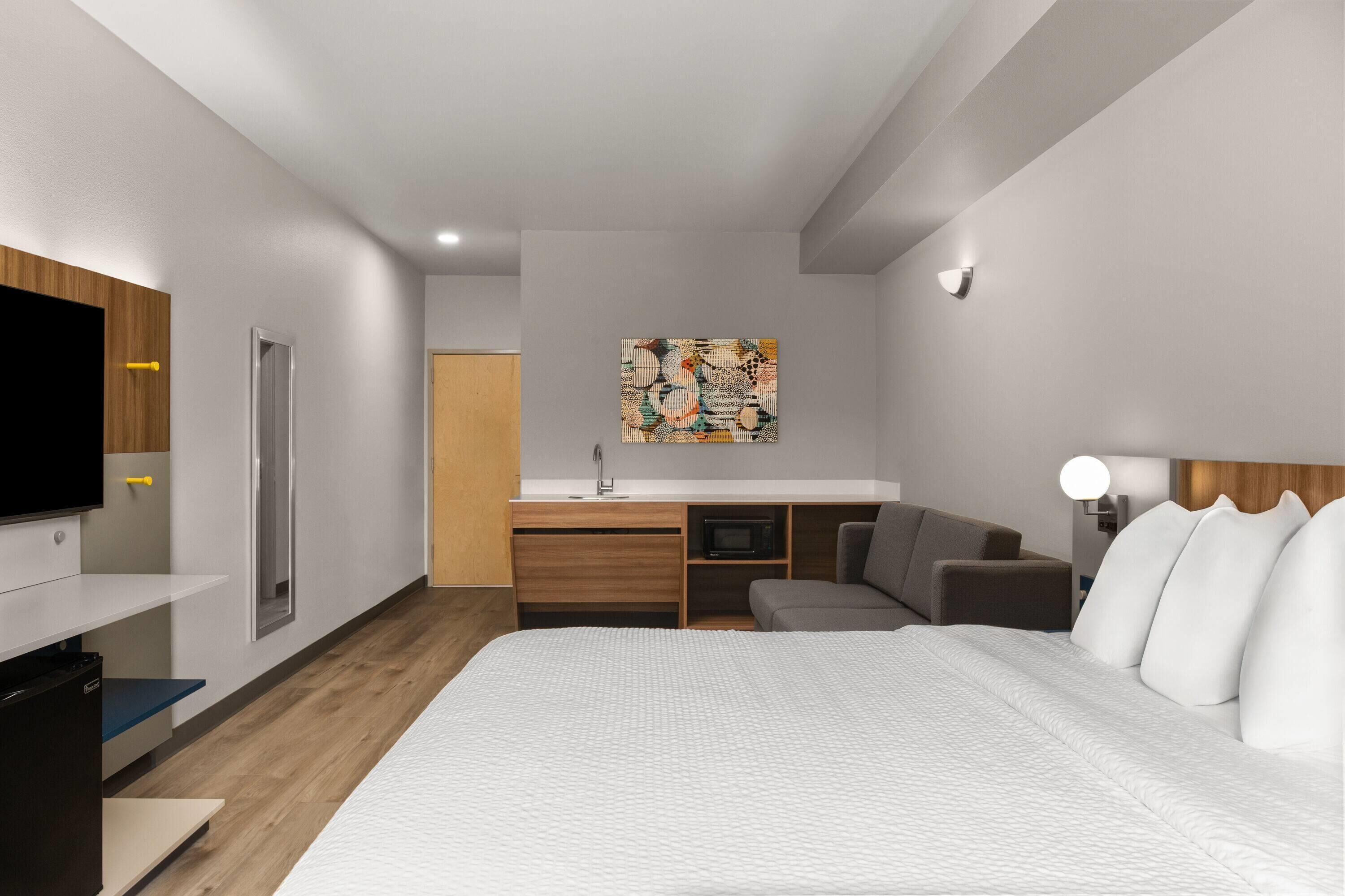 A modern hotel room with a large bed, a flat-screen TV, a desk with a microwave, a sofa, and a painting on the wall.