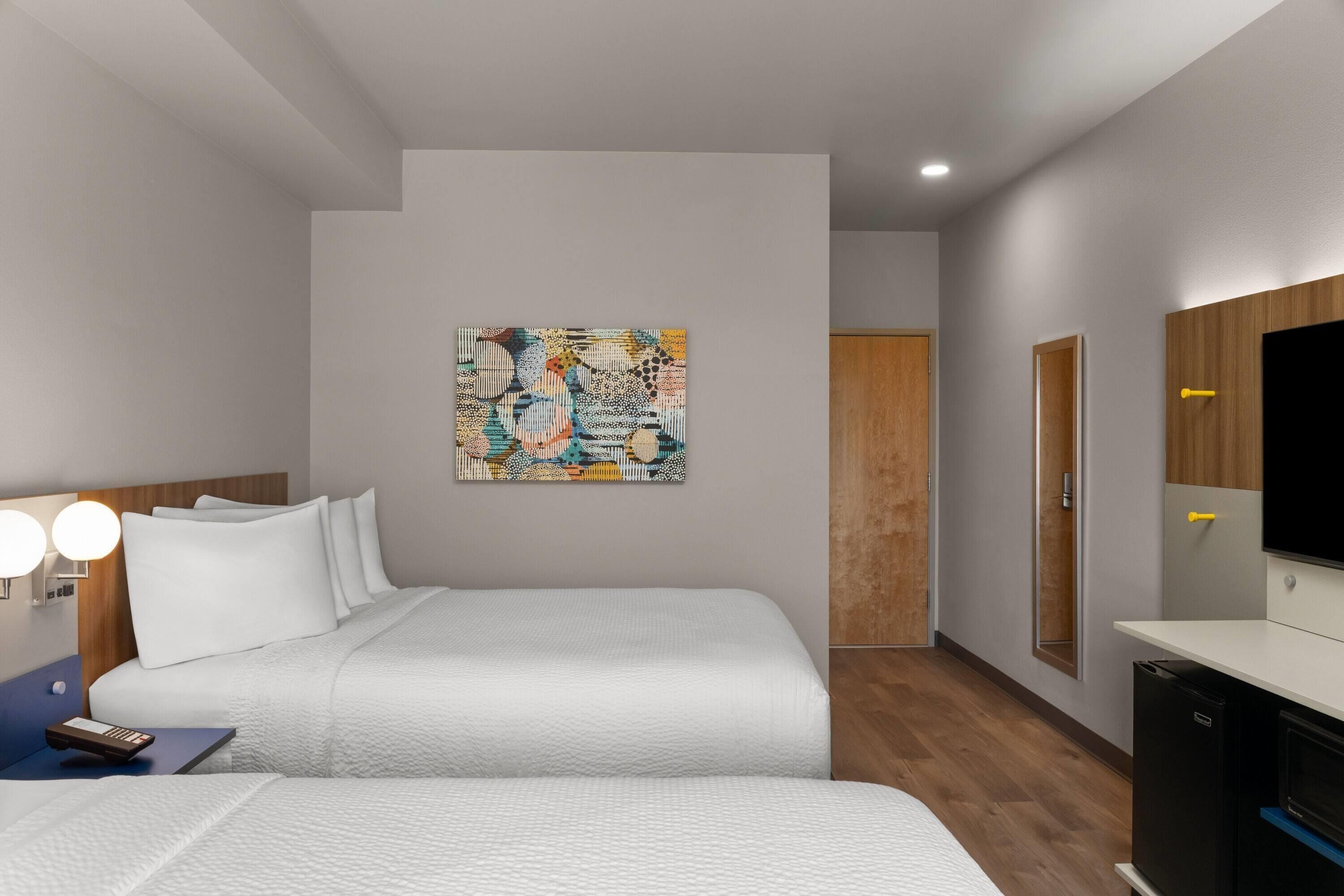 A modern hotel room with a bed, a bedside table, a lamp, a painting on the wall, and a television.