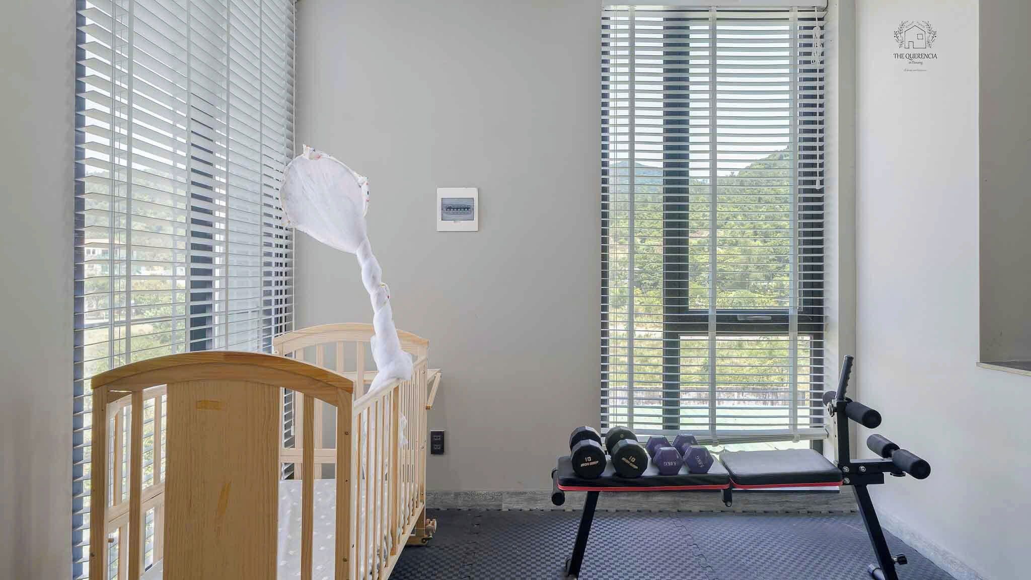 A room with a crib, a window with blinds, a wall-mounted thermostat, and a bench with dumbbells.