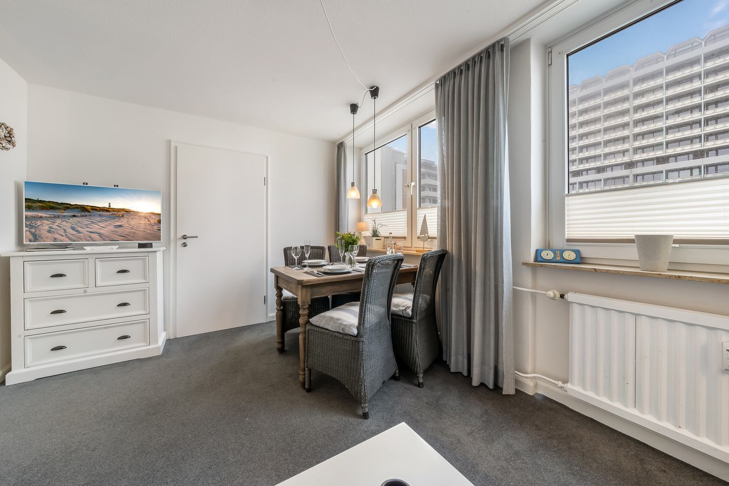 Habitación de hotel con comedor, televisor, cómoda blanca, ventana con cortinas y vista a un edificio.