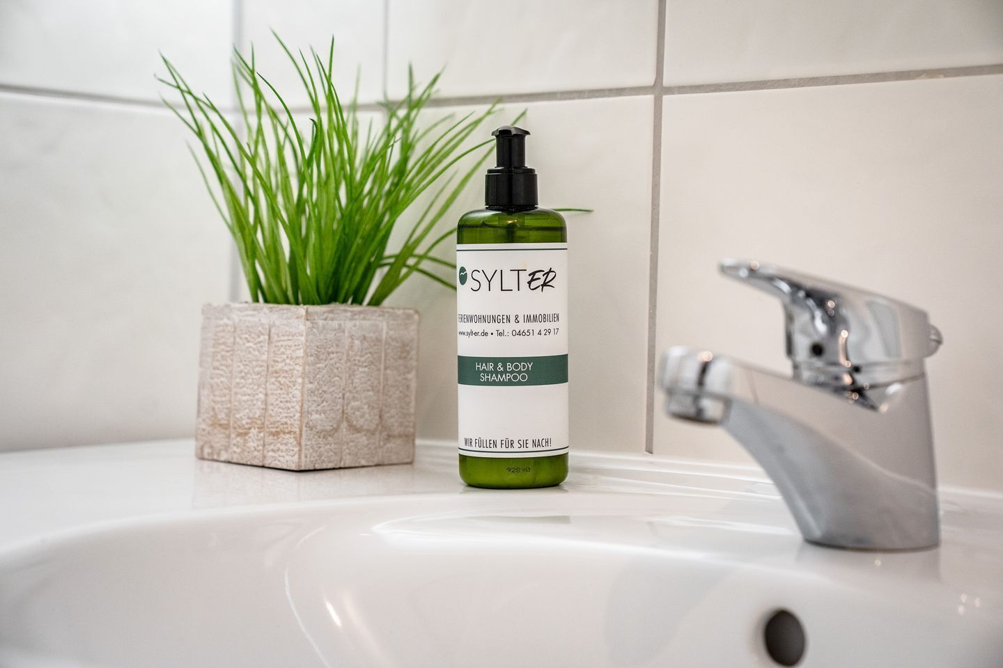 A bathroom sink with a green bottle of SYLTER hair and body shampoo, a potted plant, and a modern faucet.