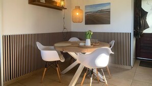 A dining area with a round wooden table, white chairs, a hanging light fixture, a wall-mounted shelf with a decorative item, and a framed picture on the wall. - Forest cottage by the sea (Sint Maartensvlotbrug)