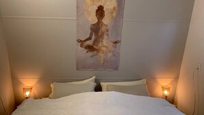 A bedroom with a bed, two bedside lamps, and a wall-mounted painting of a meditating figure with a full moon. - Forest cottage by the sea (Sint Maartensvlotbrug)
