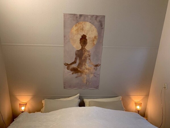 A bedroom with a bed, two bedside lamps, and a wall-mounted painting of a meditating figure with a full moon.