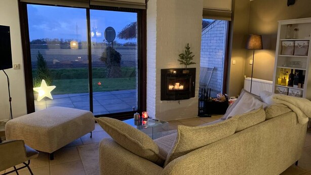 A living room with a sofa, ottoman, and a fireplace. There is a large window with a view of the outside. - Forest cottage by the sea (Sint Maartensvlotbrug)