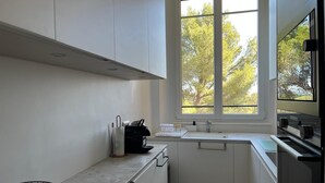 A modern kitchen with white cabinets, a window with a view of trees, a sink, and a stove. - Seasonal rental saint Raphael Valescure 80m² 4 beds (Saint-Raphaël)