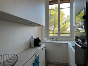 A modern kitchen with white cabinets, a window with a view of trees, a sink, and a stove. - Seasonal rental saint Raphael Valescure 80m² 4 beds (Saint-Raphaël)