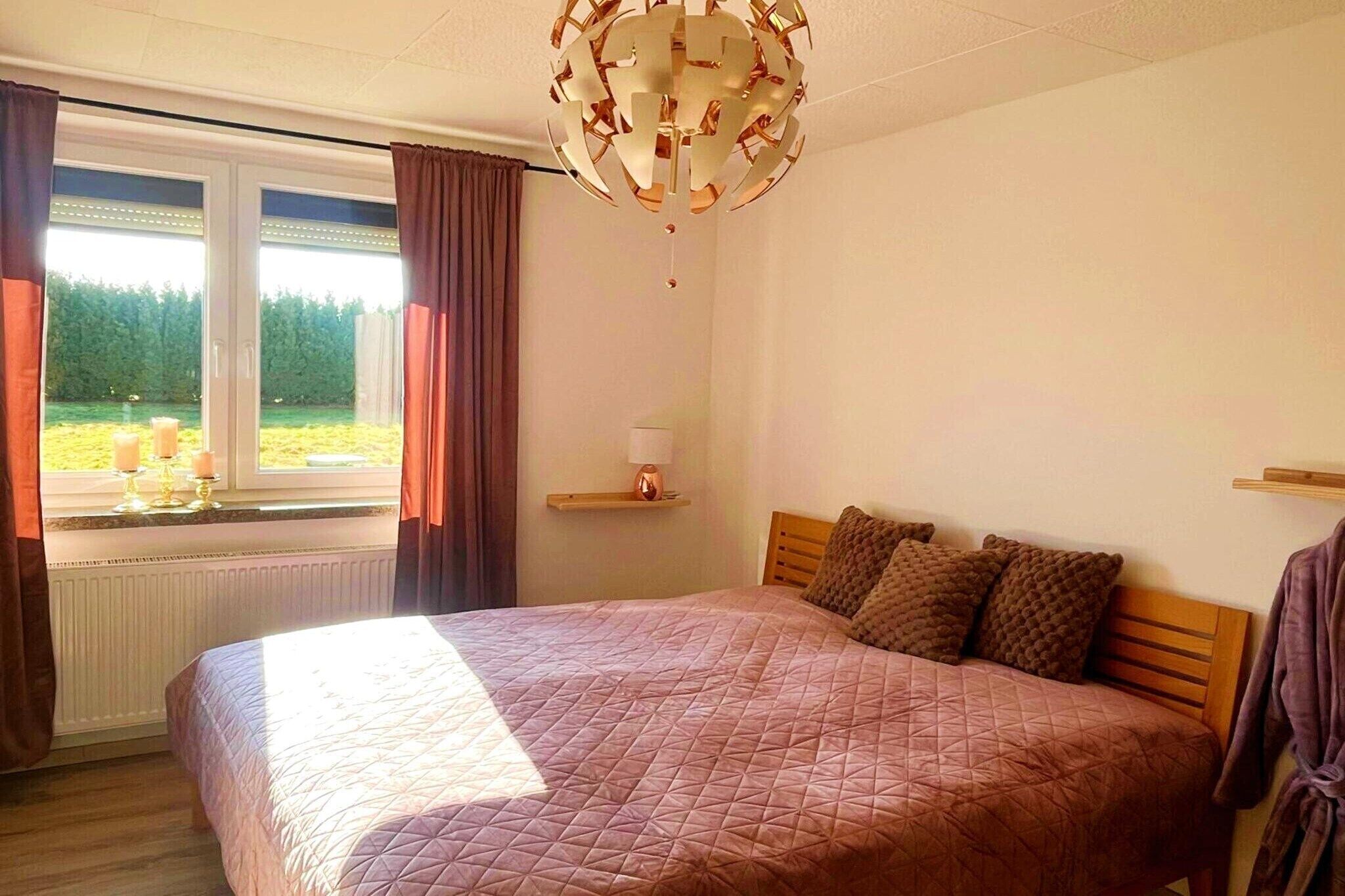 Ein Schlafzimmer mit einem großen Bett, einem Fenster mit Vorhängen, einer Chandelier und Blick ins Grüne.