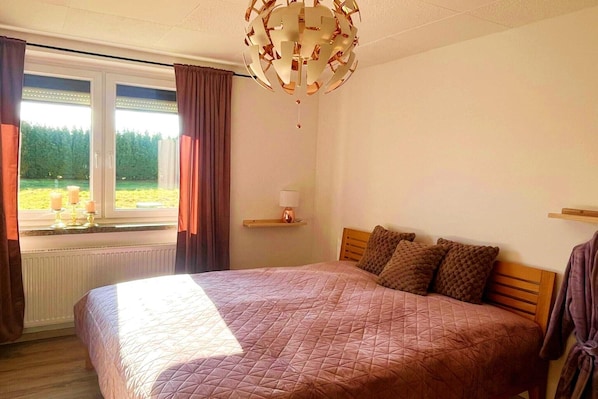 A bedroom with a large bed, a window with curtains, a chandelier, and a view of greenery outside.