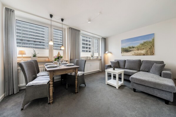 Ein modernes Wohnzimmer mit einem grauen Sofa, einem Esstisch mit Stühlen und einem großen Fenster mit Vorhängen.