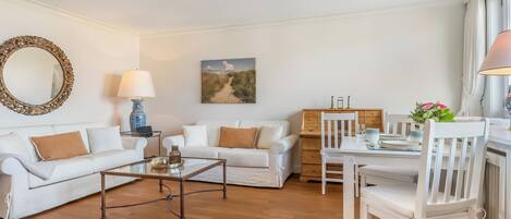A living room with a white sofa, a coffee table, a dining table with chairs, a lamp, a painting, and a mirror.