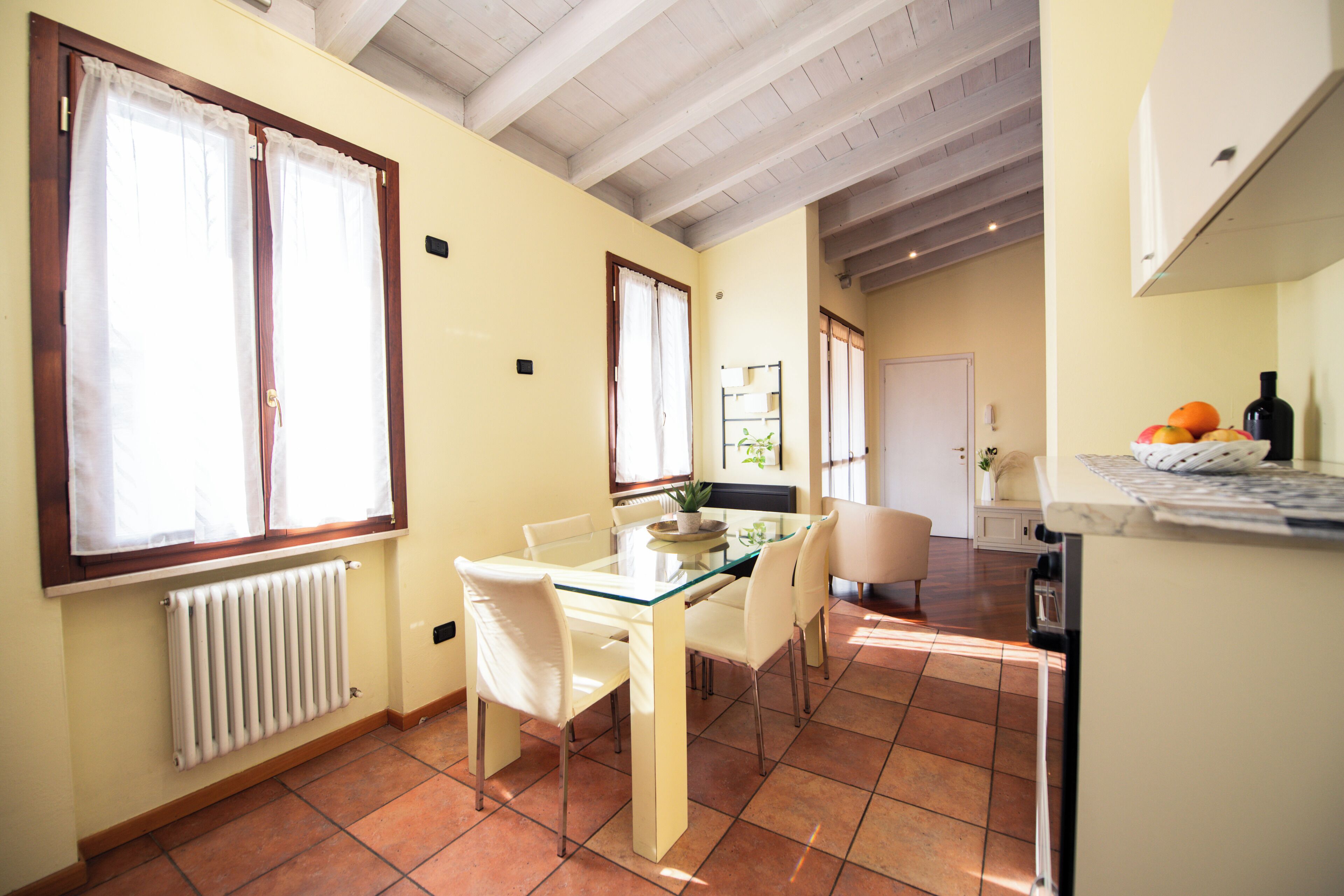 A dining area with a glass table and white chairs, a radiator, and a window with curtains.