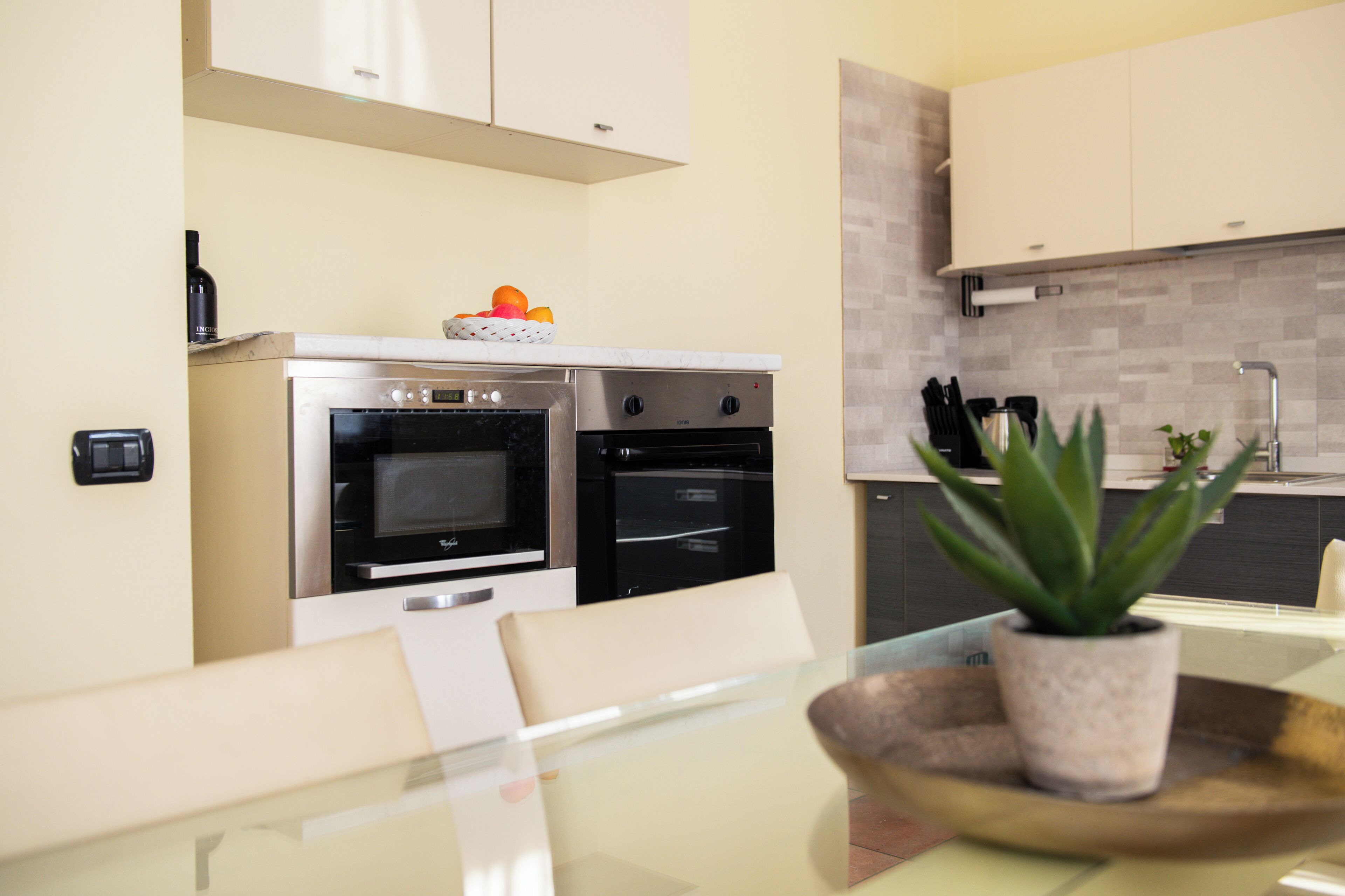 Una cocina moderna con dos hornos, un microondas y un lavamanos. Hay una planta en maceta sobre la encimera y una botella de vino en la estantería.