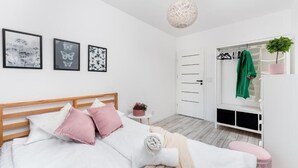 A modern bedroom with a wooden bed, white bedding, and pink pillows. There are three framed pictures on the wall, a small side table with a plant, and a window with a green towel hanging. - Malila Apartments Konopnickiej 63E (Kraków)