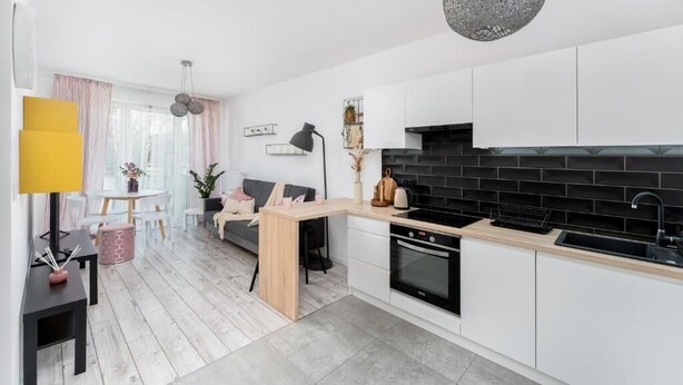 A modern kitchen with white cabinets, a black backsplash, and a wooden countertop. There is a dining area with a white table and chairs, and a sofa with a pink cushion. - Malila Apartments Konopnickiej 63E (Kraków)