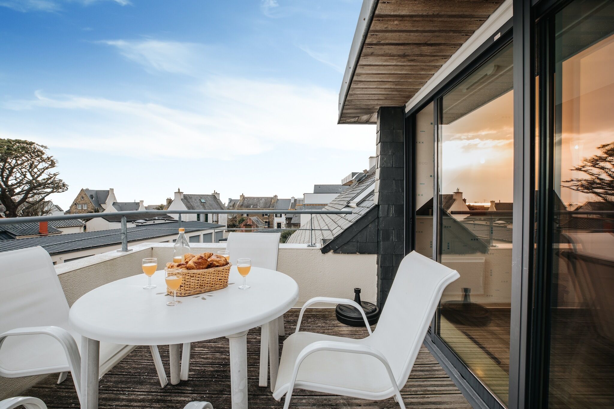 A rooftop terrace with a round table, white chairs, and a basket of bread and drinks.