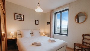 A neatly made bed with white linens and two folded towels, a wooden chair, a bedside table with a lamp, a framed picture on the wall, and a large window with a view. - Seaview Duplex with Terrace in Le Conquet (Le Conquet)
