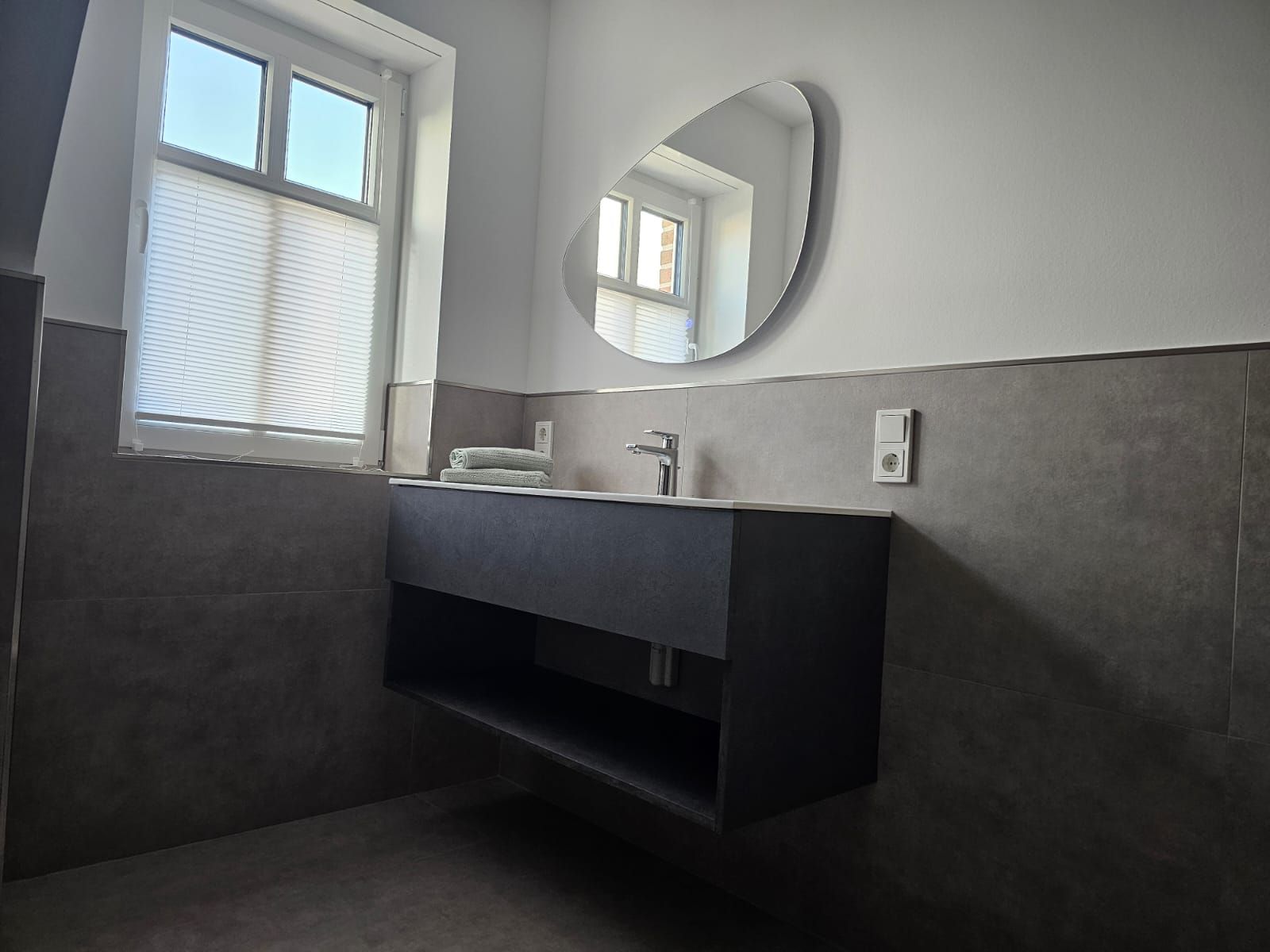 A modern bathroom with a dark grey vanity, a round mirror, and a window with blinds.