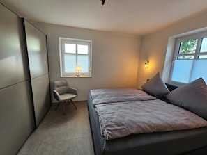 A bedroom with a bed, a chair, a window with blinds, and a wall-mounted lamp.