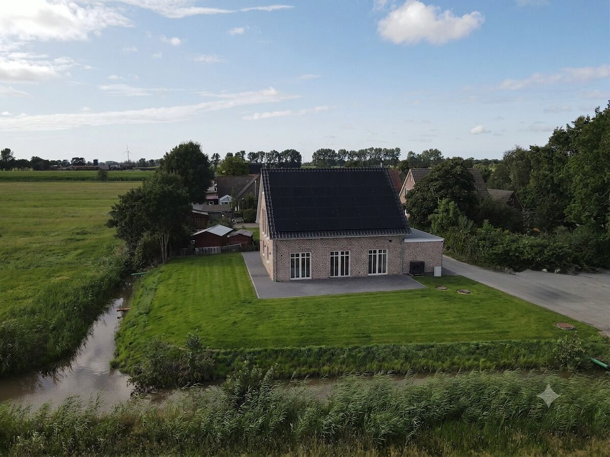 Een laag gebouw van baksteen met een schuin dak, omgeven door groene velden en een kleine beek.