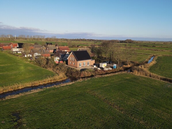 A rural landscape with a house, a small stream, and wind turbines in the distance. - Vacation home 53° North - new building 2026 (Hinte)