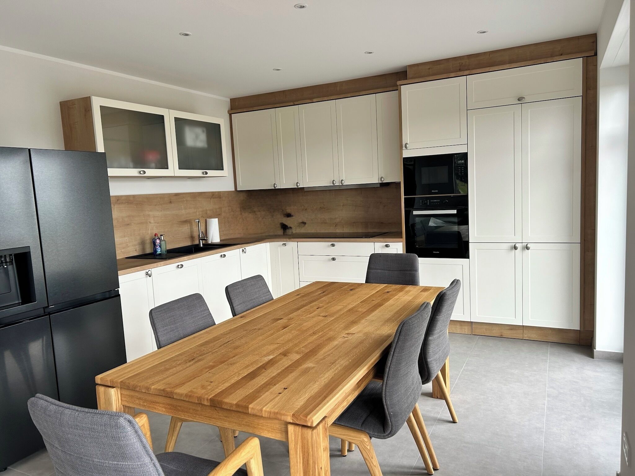 Een moderne keuken met een houten eettafel, grijze stoelen, witte kasten en inbouwapparatuur.
