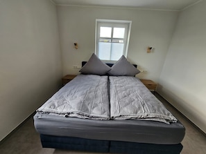 A narrow room with a bed, two bedside tables, and a window with blinds.