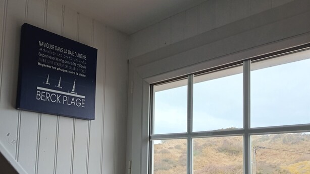 Ein Zimmer mit großem Fenster und Blick auf eine hügelige Landschaft. Ein Schild an der Wand trägt die Aufschriften 'NAVIGUER DANS LA BAIE D'AUTHIE' und 'BERCK PLAGE'.
