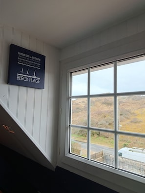 Une pièce dotée d’une grande fenêtre donnant sur un paysage vallonné. Une pancarte sur le mur indique : « NAVIGUER DANS LA BAIE D’AUTHIE » et « BERCK PLAGE ».